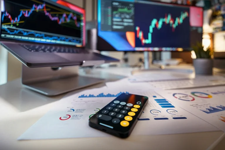 A desk with a calculator and printed charts in front of a computer screen showing stock market graphs