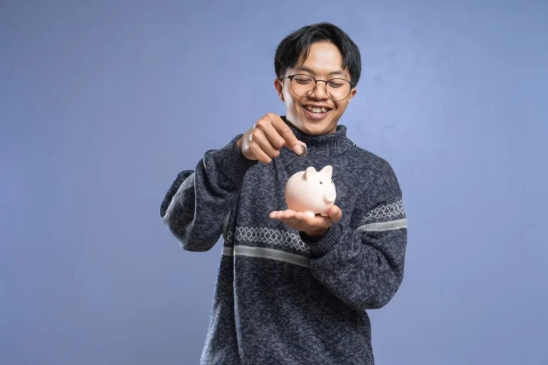 Smiling young man putting money into a piggy bank representing good money habits and wealth building