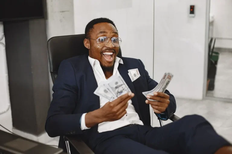 A smiling man sitting in an office chair holding cash representing financial freedom and success