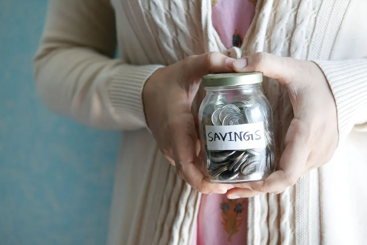 A person holding a glass jar labeled Savings filled with coins for monthly money saving