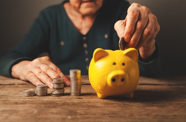 An older person placing a coin into a yellow piggy bank next to coin stacks representing retirement savings