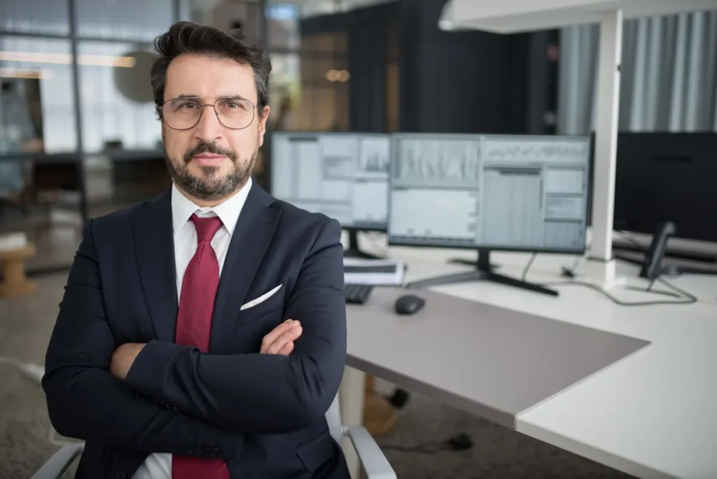 A professional businessman in a suit sitting at a desk in a modern office and represent Who Is a Businessman