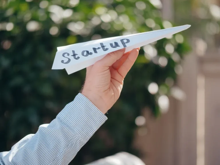 Hand holding a paper airplane with startup text representing Business Startup Costs.