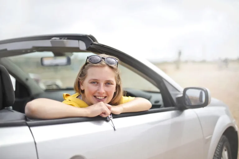 Young woman smiling while leaning out of a convertible car window, illustrating How Does Car Insurance Work in the USA? for drivers.